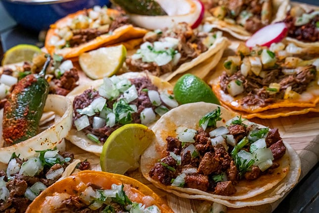 Multiple  mini tacos are laid out on a table. You can see the close up on the meats, onions, and shells. There are also slices of lemons on the table. 