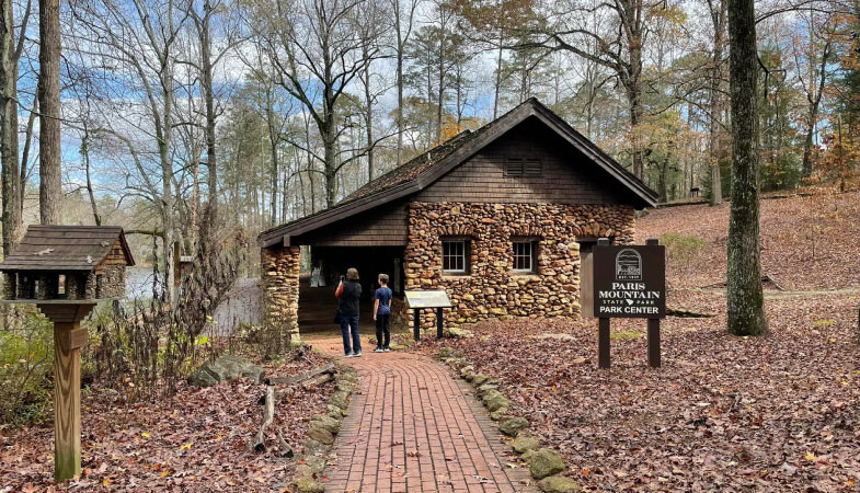 A mother and son are exploring Paris Mountain State Park in Travelers Rest, South Carolina. The Park Center is made of stones and wood siding, there is a brick path lined with natural stones, and the grounds are covered in fallen leaves.