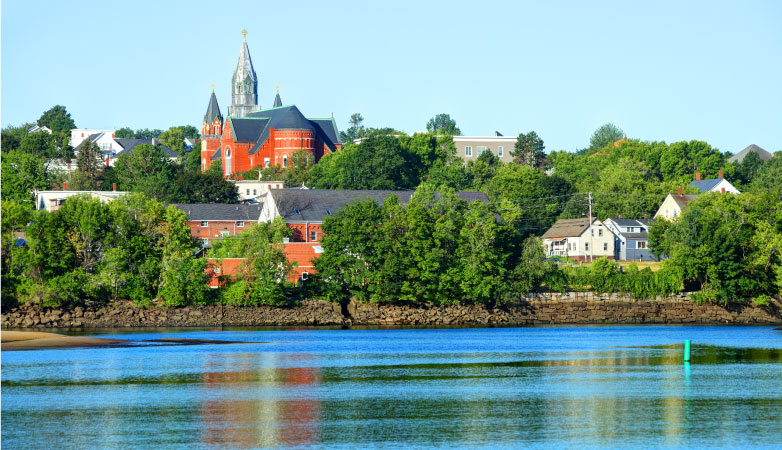 View of Biddeford, Maine, from the water. Various homes, brick buildings, and a church are seen poking out from between lush, green trees in the summer.