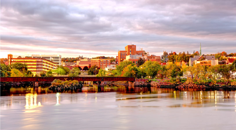 View of Bangor, Maine, from the water just before sunset. The sky is filled with fluffy clouds and the city’s buildings reflect warm hues from the setting sun. 