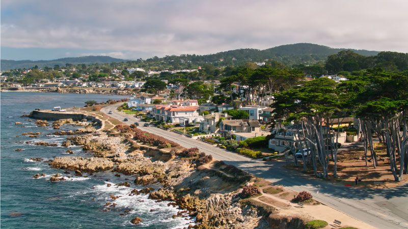Aerial view of Pacific Grove, California, a small coastal town in Monterey County. A road runs along the rocky coast and there are low tree-covered hills in the background.