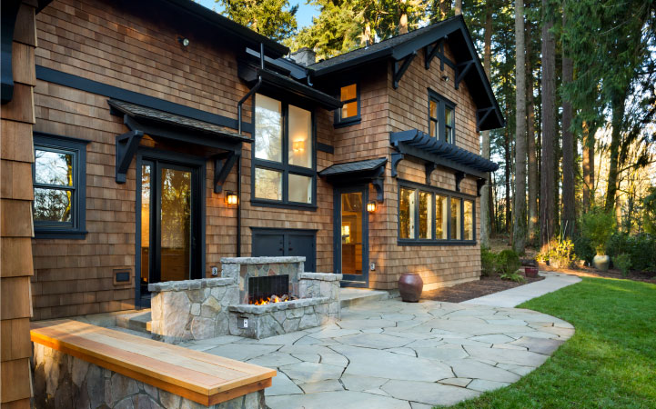 A brown shingled house with a backyard stone patio. The patio has a built-in outdoor fireplace and bench seating.