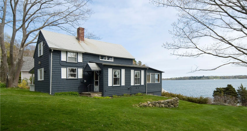 A blue shingled waterfront home in Yarmouth, Maine. The home is two-stories with a brick chimney and there’s a short stone wall in the lush yard.