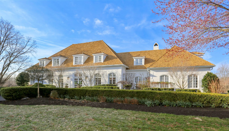 A large, single-family home, surrounded by manicured hedges in North Potomac, Maryland. The roof is tiled in a light beige and the exterior appears to be stucco, painted in a light pink. Over a dozen windows let natural light into the front of the home. 