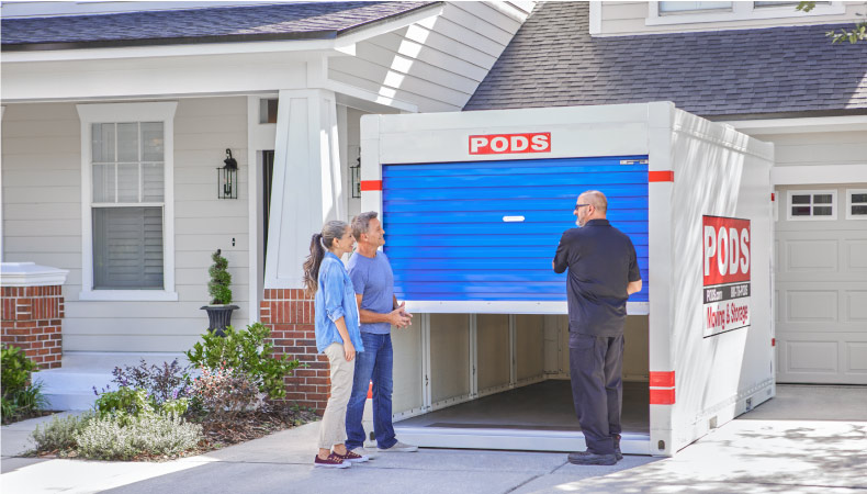 A PODS driver is showing a couple their PODS portable storage container in the driveway of their home. They’re going to use it during their bedroom remodel to store furniture, tools, and other equipment.