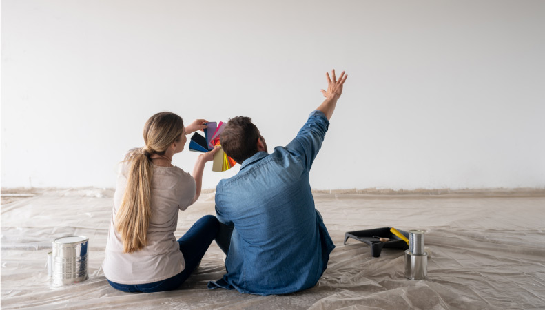 A couple is sitting on a plastic sheet on the floor of their bedroom, holding up color swatches and imagining what the wall would look like in different colors. There are paint cans on the floor beside them.