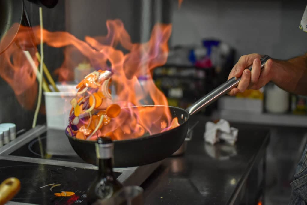 A frying pan with food and a large flame within it over a ghost kitchen stove