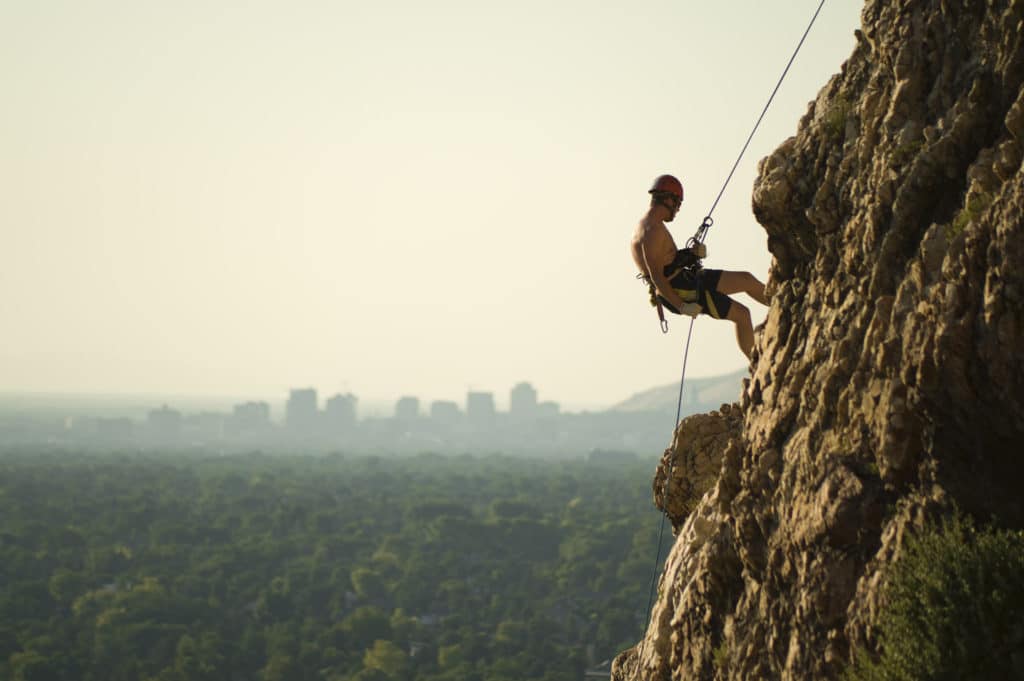 Rock climber hanging by harness at side of mountain with Salt Lake City skyline in distance