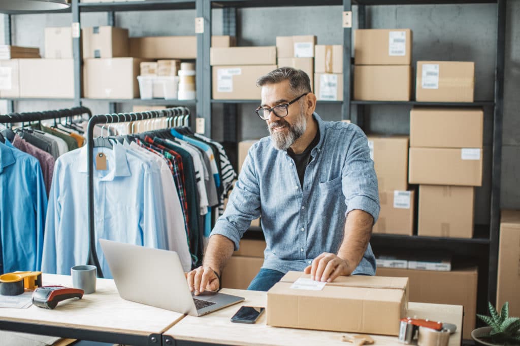 Male retail employee in backroom filled with boxes managing e-commerce orders on a laptop