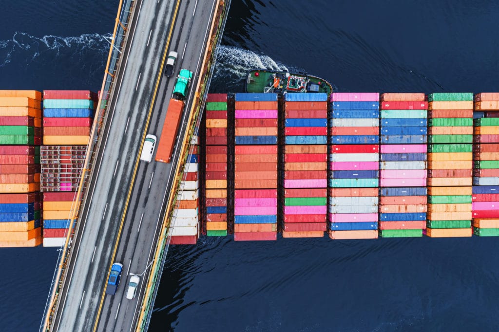 Aerial view of rows of shipping containers on a boat passing under a highway bridge