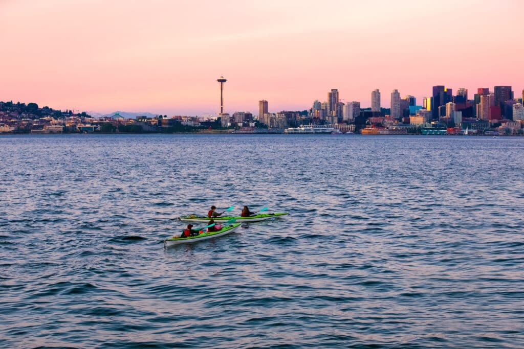 Four people in kayaks in Puget Sound waters with skyline and cityscape of Seattle in background at sunset