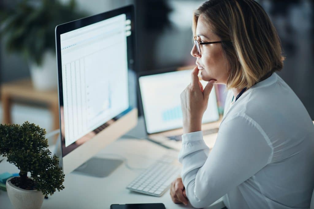 Middle-aged office woman in deep thought while looking at her desktop computer and laptop