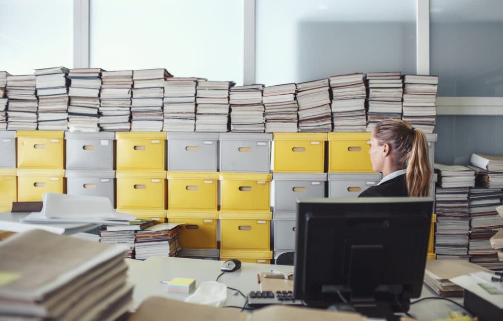 Young female office worker looking at stacks of boxes and paper clutter behind her desk