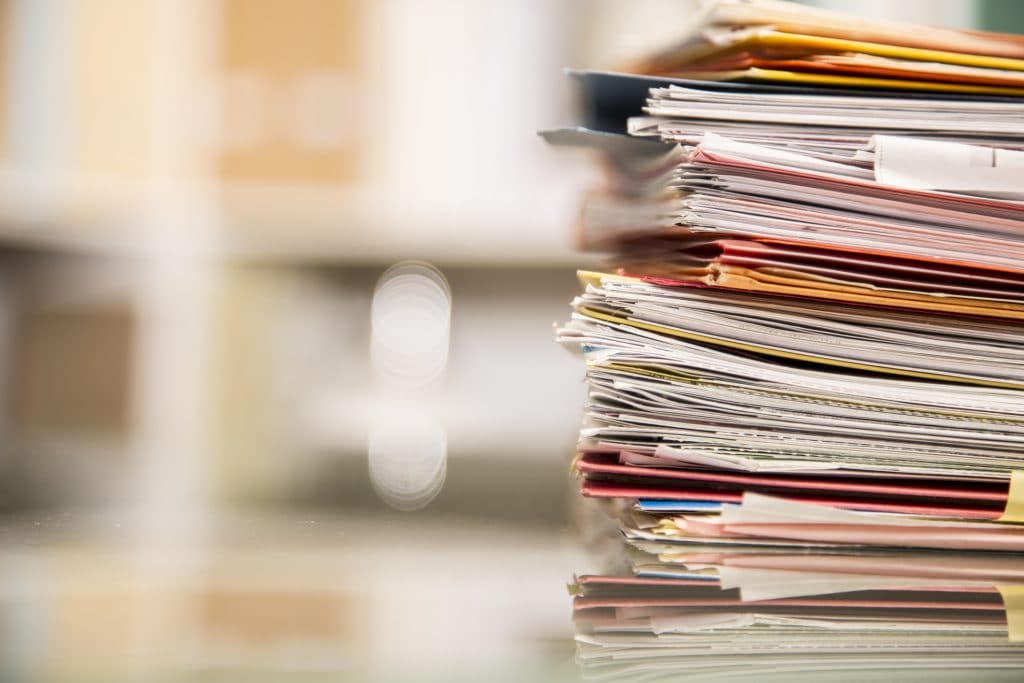 Large stack of file folders, documents, paperwork piled on glass top desk in office. Bookshelves in background.