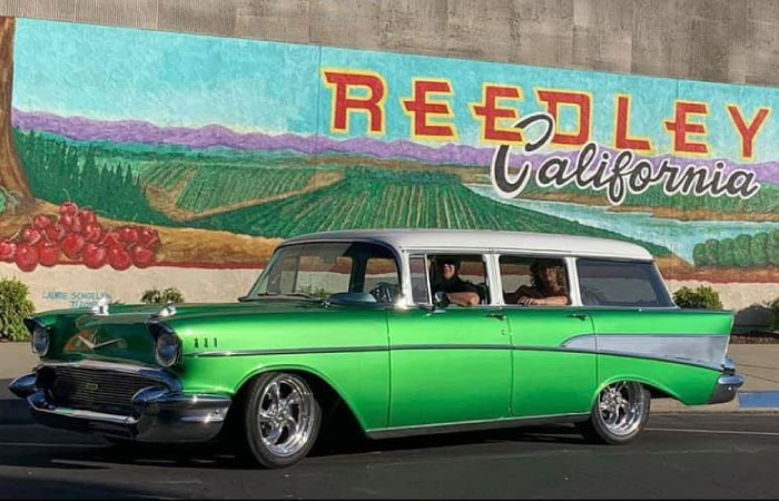 Two people are parked in a classic car from the 1950s in front of an agriculture-themed mural in Reedley California