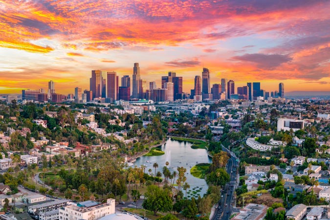 Aerial sunset view of Los Angeles, California, featuring a vivid sky, city skyscrapers, and a lush city park with a pond in the foreground.