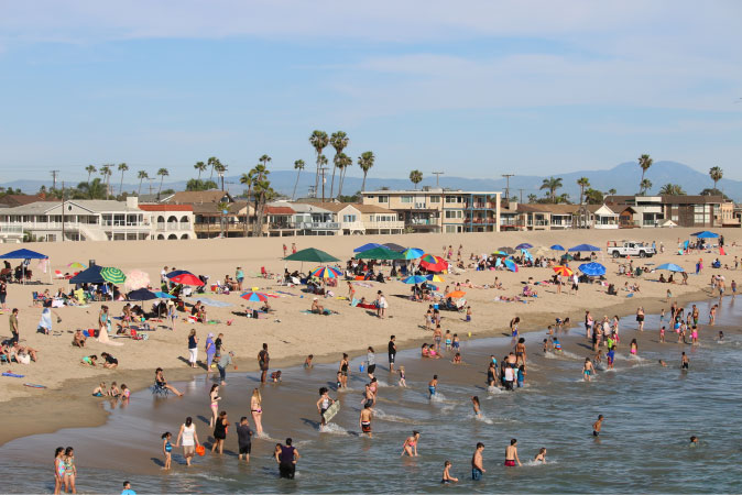 View of a crowded beach in Seal Beach, California, on a summer day, featuring hundreds of beachgoers, colorful umbrellas, and a mountain range in the distance.