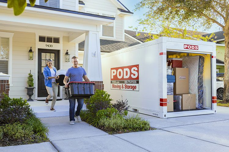 A mature couple carries items to load into their nearly full PODS container that sits in their driveway. The container is white and reads “PODS Moving & Storage” on the side.