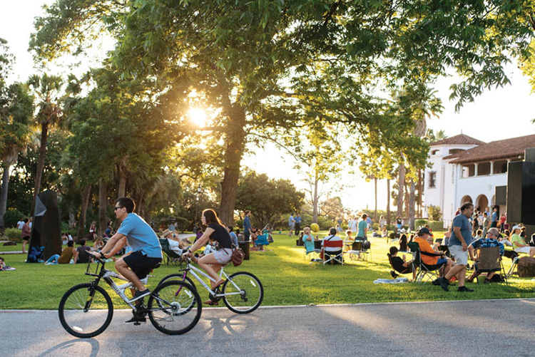 A crowd of people sit on the grass and ride bikes at a park in Alamo Heights. There is sunlight coming through a tree and a white building in the background. 