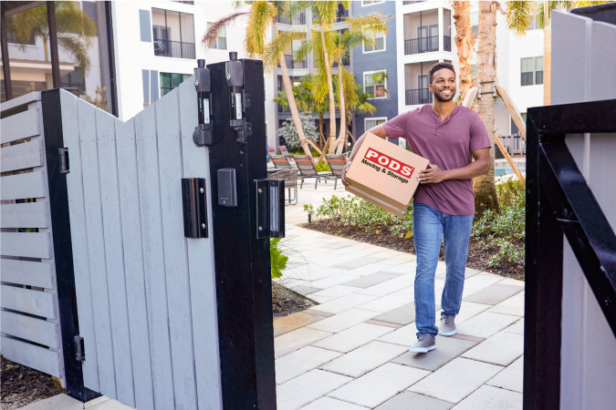 A young man is smiling as he carries a moving box with PODS branding through his apartment complex. 