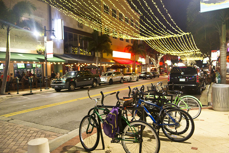 A view of Clematis Street in downtown West Palm Beach. Bikes are the main focus of the image, with string lights above them. On the other side of the street, people are drinking at a bar. 