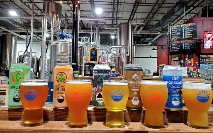 Five glasses of cold craft beer are lined up on a wooden ledge in a brewery in Raleigh, North Carolina. Behind the glasses of beer are the individual beer cans.
