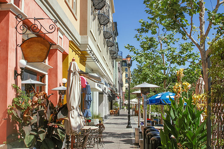 A row of colorful stores in downtown San Jose, California, during the day. There is greenery and chairs and tables outside of a cafe. 