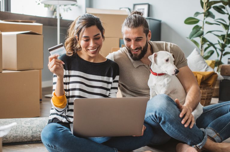 A couple and their dog are sitting on the floor near stacks of moving boxes, estimating the cost of moving to their new home