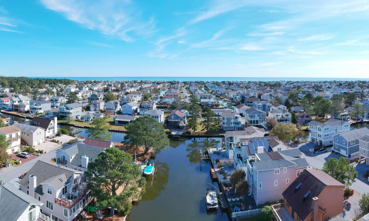 Aerial view of a neighborhood filled with canal-facing homes in Bethany Beach, Delaware. Some of the homes feature large trees in their backyards, and nearly all of them have a private dock on the canal. In the distance, beneath a blue sky, the coast is visible on the horizon.