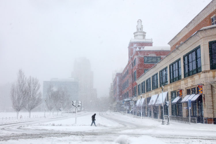 A person walks through downtown Providence, Rhode Island, during a nor’easter snowstorm. The streets are covered in a hefty layer of snow, and the buildings in the distance are obscured by the flurry.