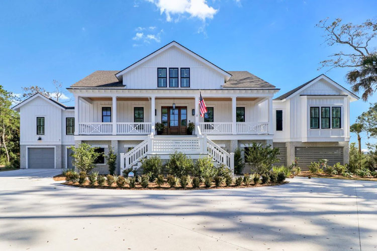 A large residential home in Dutch Island, Georgia, outside of Savannah. The home features a dual staircase leading up to a large covered porch and entrance, a two-car garage, and a white exterior. 