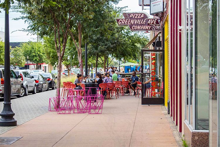 The neighborhood of Lower Greenville in Dallas, Texas. There are colorful tables and chairs outside restaurants for people to sit at. There is a sign that reads “Greenville Ave Pizza Company.” 
