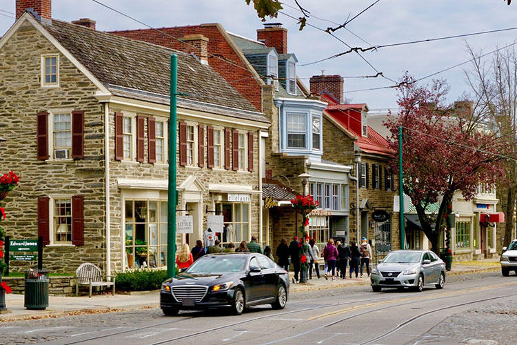 A view of downtown Chestnut Hill in Philadelphia, Pennsylvania. There are stone buildings and a group of people walking on the sidewalk. Cars are passing by. 