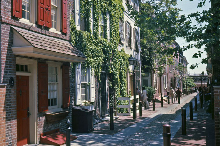 Brick row homes in the neighborhood of Queen Village in Philadelphia, Pennsylvania. There is moss covering one building and two couples walking along the sidewalk. 
