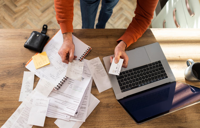 A man is hunched over his laptop and various financial papers as he works to get his finances in check.