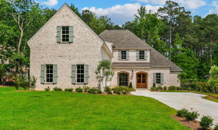 A large new construction home made of light-colored brick and set against a forested backdrop. The home is two stories tall and is set on a large lot in Flowood, Mississippi.