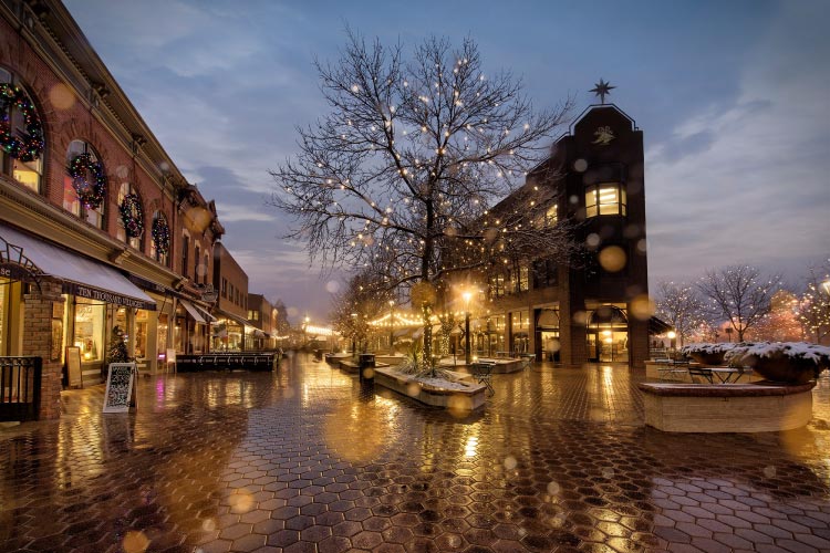 Old Town Square in Fort Collins, Colorado, is lit with Christmas lights. The hexagonal pavers are glistening in the rain, and a little bit of snow is still resting on some potted plants.