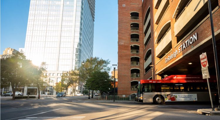 A red GoRaleigh bus is pulling out of a station in Raleigh, North Carolina, on a sunny day. The sun is reflecting brightly off a glass skyscraper in the distance, and trees are casting shadows across the city street.
