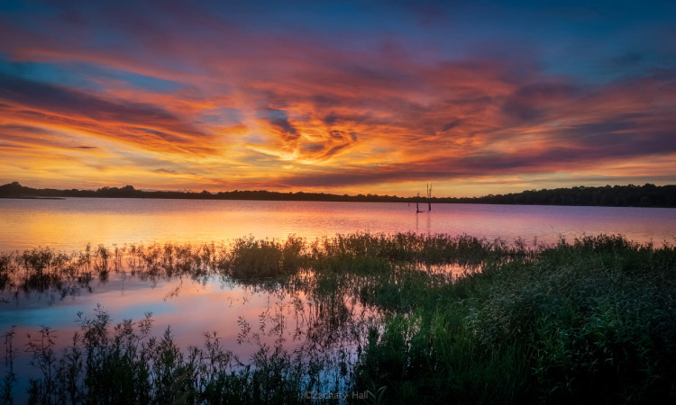 The sky is ablaze with warm hues of orange, pink, and gold, casting a mesmerizing reflection upon the calm surface of Jack Nolen Lake in Greenwood, Arkansas. The water mirrors the vibrant colors, and silhouettes of trees and distant hills are outlined against the colorful sky, adding depth to the scene.