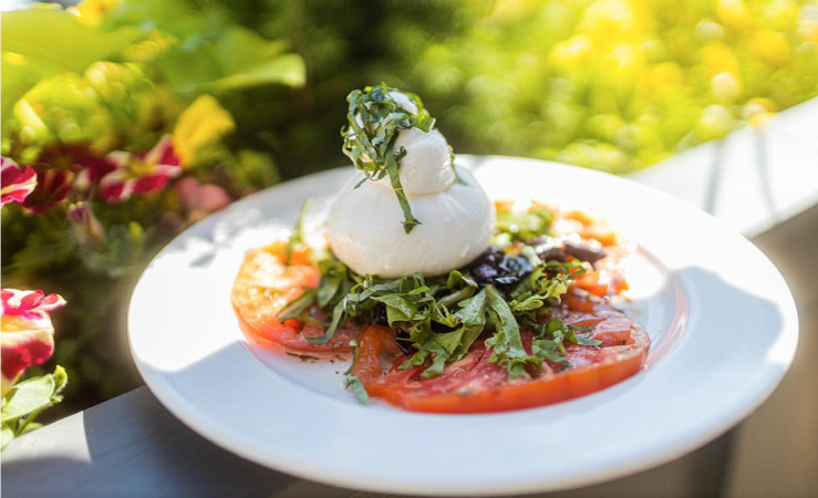 An heirloom salad with fresh tomato slices, arugula, and burrata, is arranged neatly on a white plate outside. 