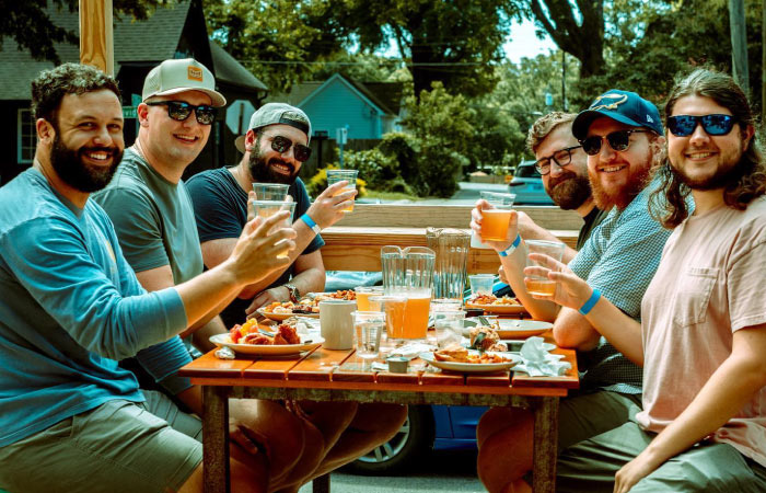 Six men smile and hold up their craft beers for a photo as they enjoy brunch at Heist Brewery in Charlotte, North Carolina. Brewery hopping is one of the many fun things to do in Charlotte, NC.