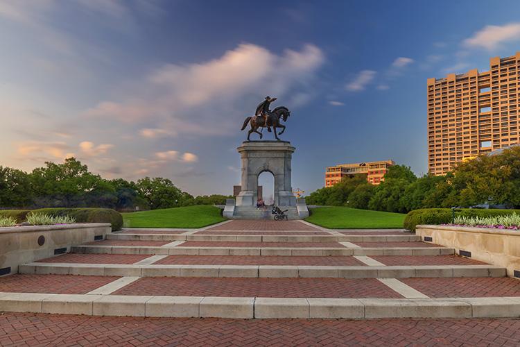An image of a monument of a person on a horse in Hermann Park in Houston, Texas. It is dusk, and there are lines of bricks for a path with buildings in the background. 