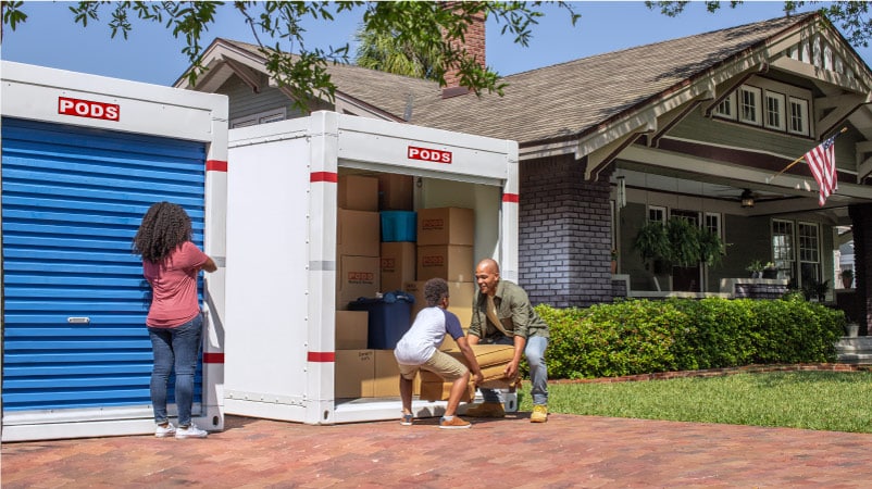 A man and his son lift a piece of furniture into a PODS portable storage container in their driveway as his wife looks on. The container is already nearly filled with neatly stacked cardboard boxes.