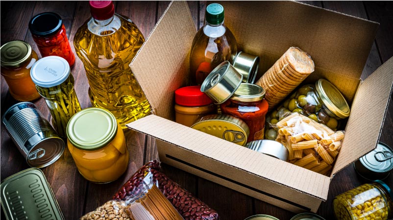 Perishable food items, including jarred and canned foods, dry pasta, crackers, and cooking oils, are packed in an open cardboard box. Some items are also sitting on the table beside the box.