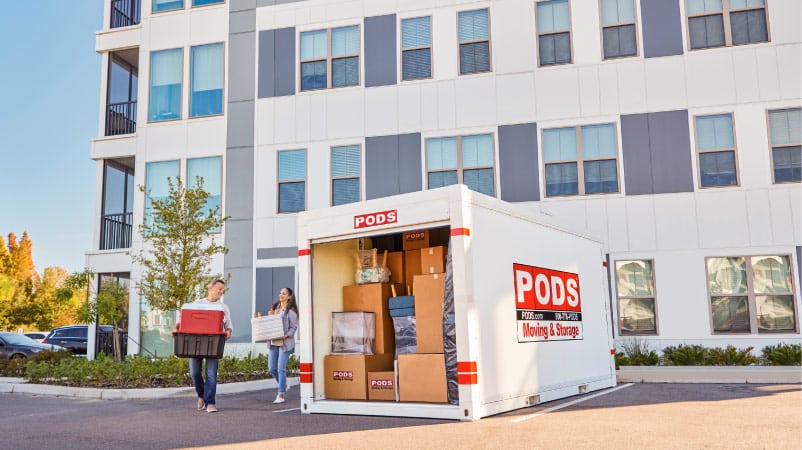 A man and woman carry a box and storage tubs to their PODS portable storage container in the parking lot of their apartment complex.