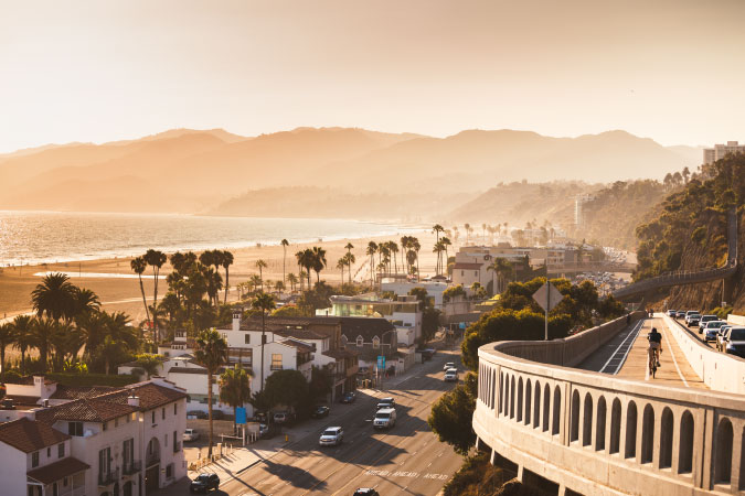 View from above of a highway in Santa Monica, California, with the Pacific Ocean and nearby mountains visible beyond the oceanfront buildings.