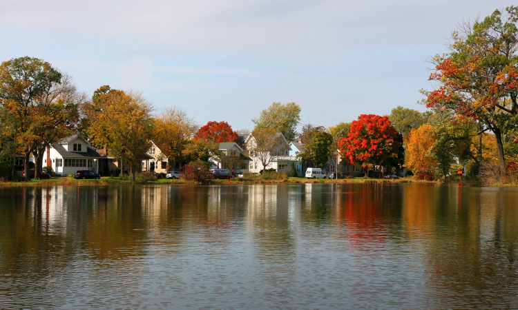 A view across the lake in Tenney Park of several large waterfront homes in the Tenney-Lapham neighborhood of Madison, Wisconsin. It’s autumn, and many of the trees around the lake have begun to change their leaves and are displaying vibrant shades of red and yellow.