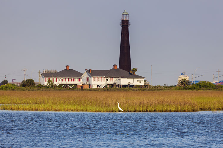 The Bolivar Island lighthouse in the daytime. An egret wades in the water in the photo’s foreground.
