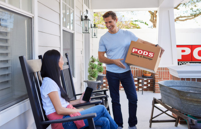 A young couple takes a break from loading their PODS portable container. The woman sits in a black rocking chair on the porch. The man is holding a brown moving box that reads “PODS Moving & Storage.” 