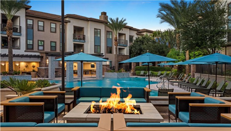 The community pool at a luxury apartment complex in Gilbert, Arizona. The pool deck features reclining chairs, umbrellas, and a gas fire pit surrounded by patio furniture.
